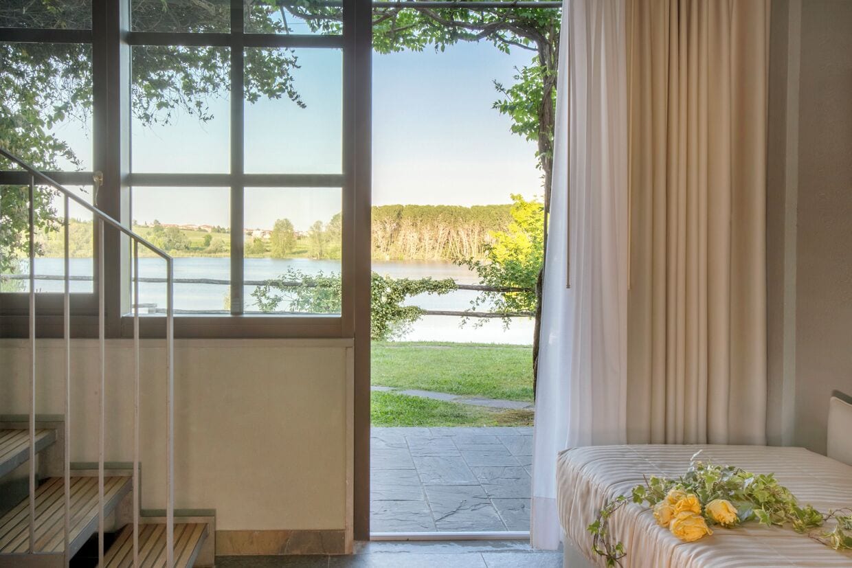 A bedroom with a view of a lake and trees, a staircase, and a bed with a floral arrangement.