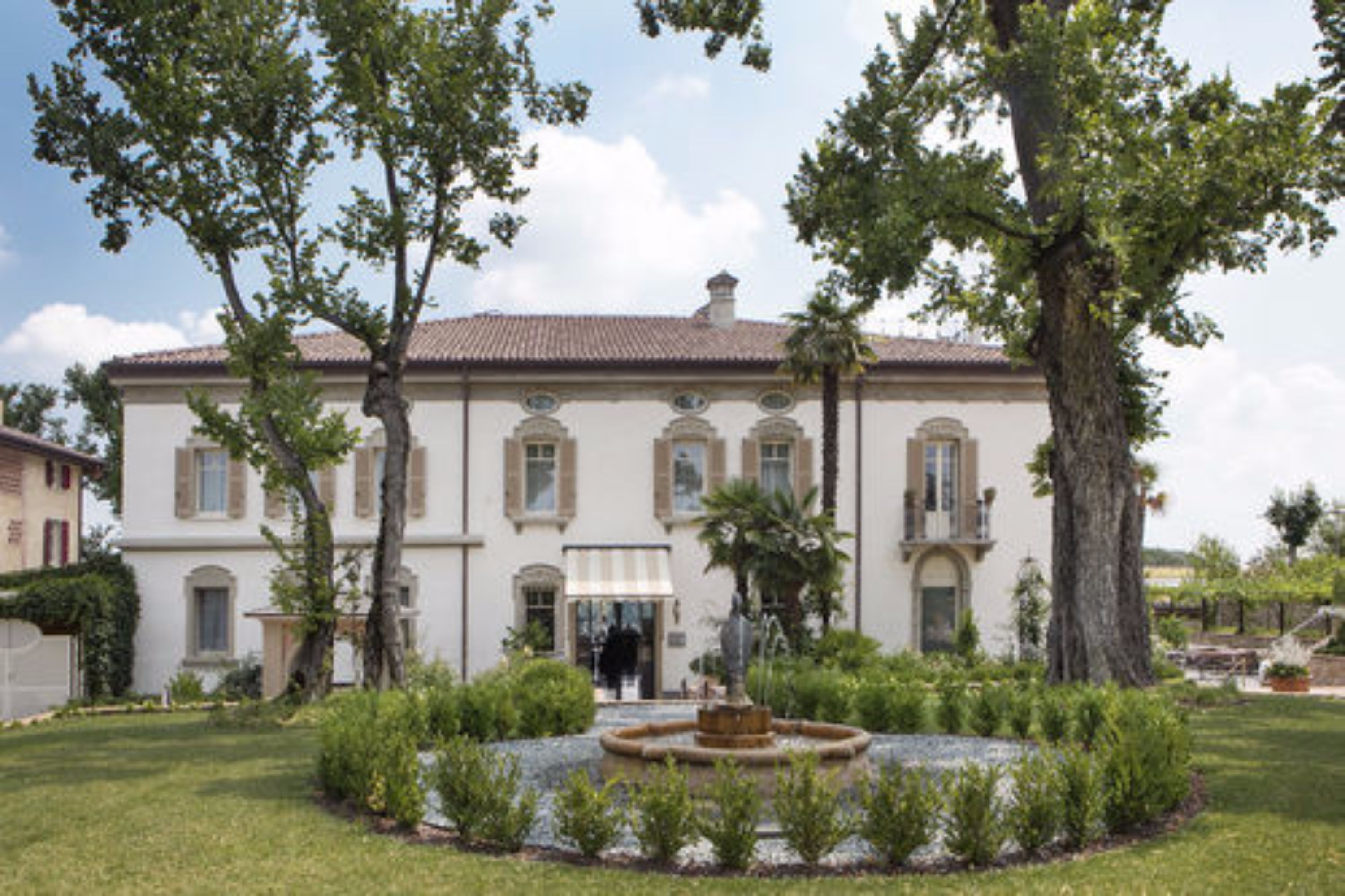 A large, white building with a tiled roof, surrounded by a well-maintained garden and a central fountain.