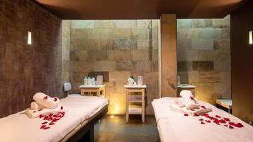 Ett spa-rum med två sängar, var och en med ett handduk och rosenblad, och en stenmur i bakgrunden.