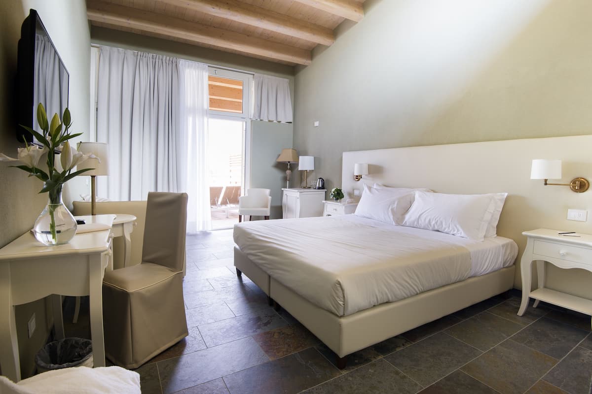 Habitación de hotel con una cama grande, un escritorio con lámpara, una silla y vistas al exterior a través de una ventana con cortinas.