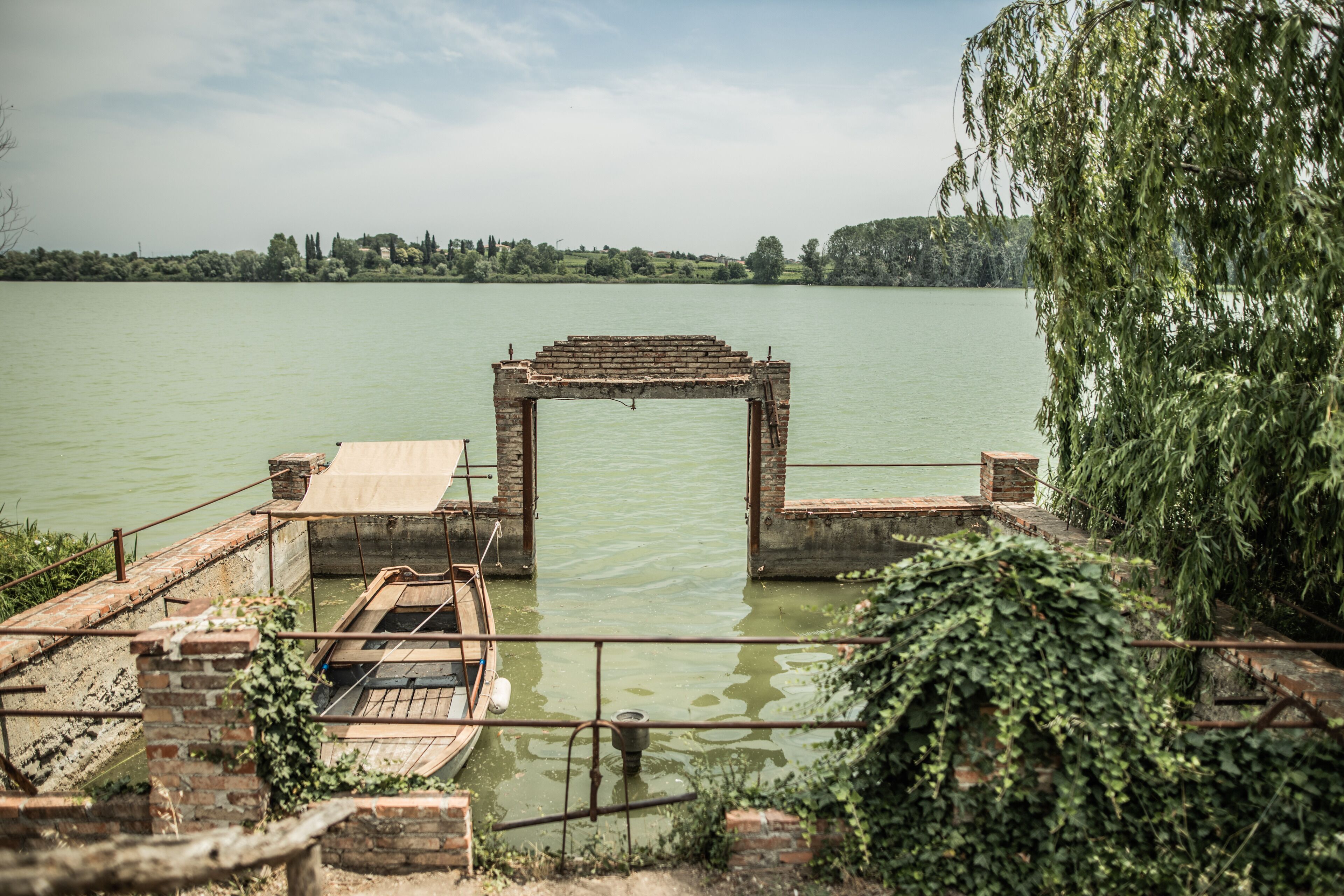 Une jetée en bois rustique, surmontée d’une arche en briques qui mène à un lac paisible, le tout entouré de verdure et dominé par des collines lointaines.