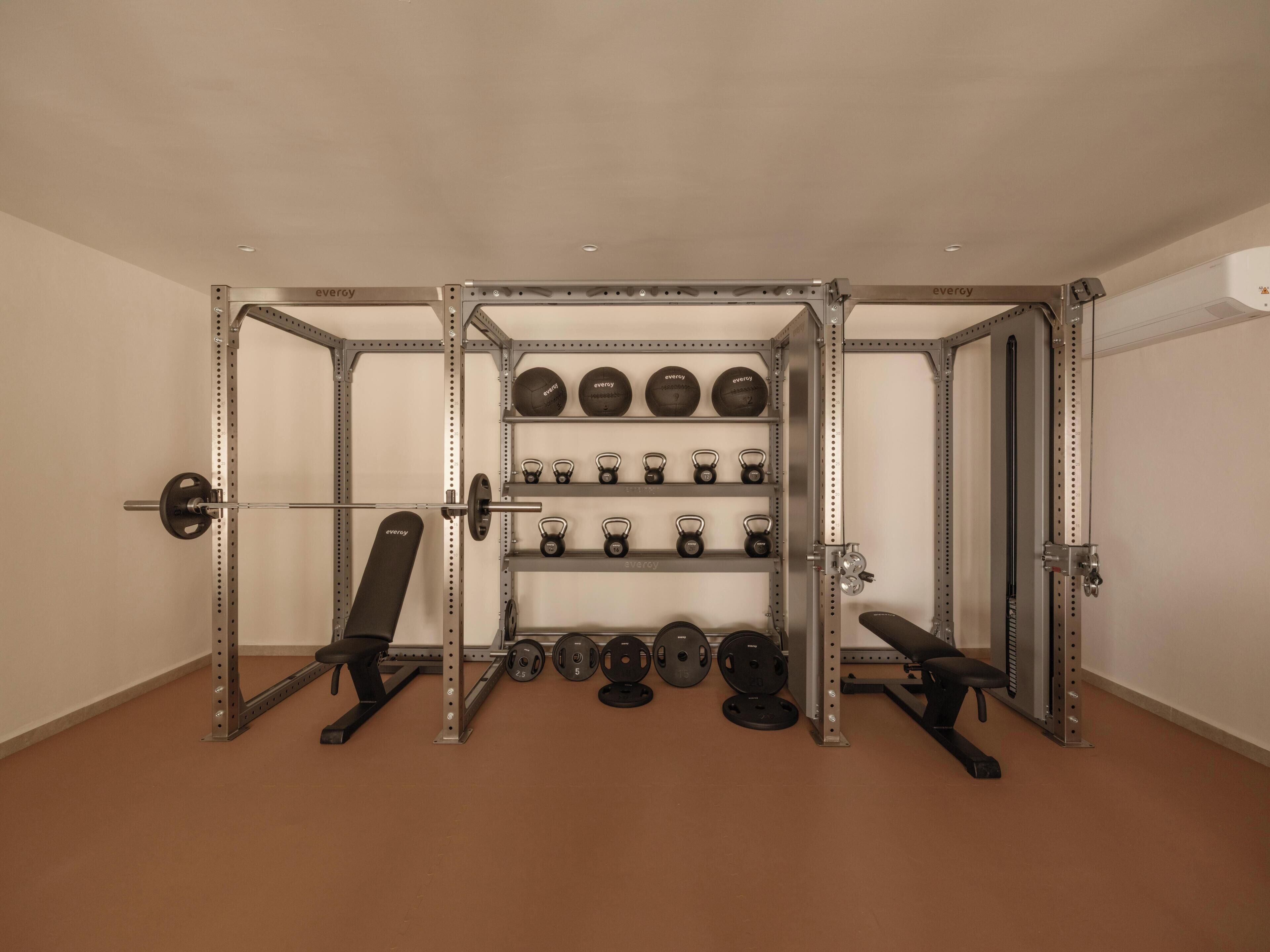 Un gimnasio con equipo de levantamiento de pesas, incluyendo una estructura para sentadillas, bancos y diferentes pesas.