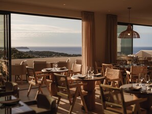 Un comedor con mesas y sillas, ventanas grandes y vista al océano.