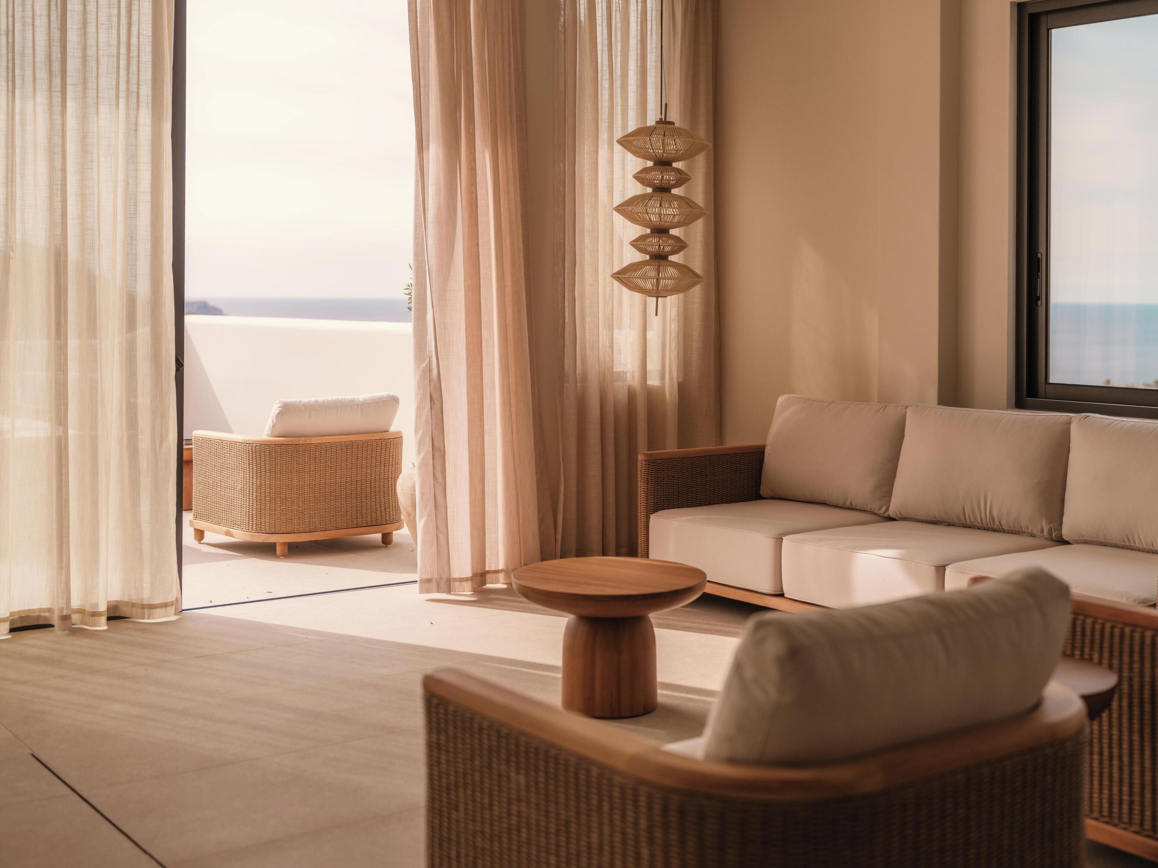 A modern living room with a sofa, a round wooden side table, a wicker chair, and a view of the sea through large windows.