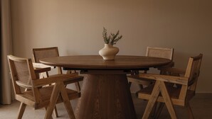 Une table ronde en bois, sur laquelle est posé un vase contenant des plantes séchées, entourée de six chaises en bois dont les dossiers sont en osier tressé.