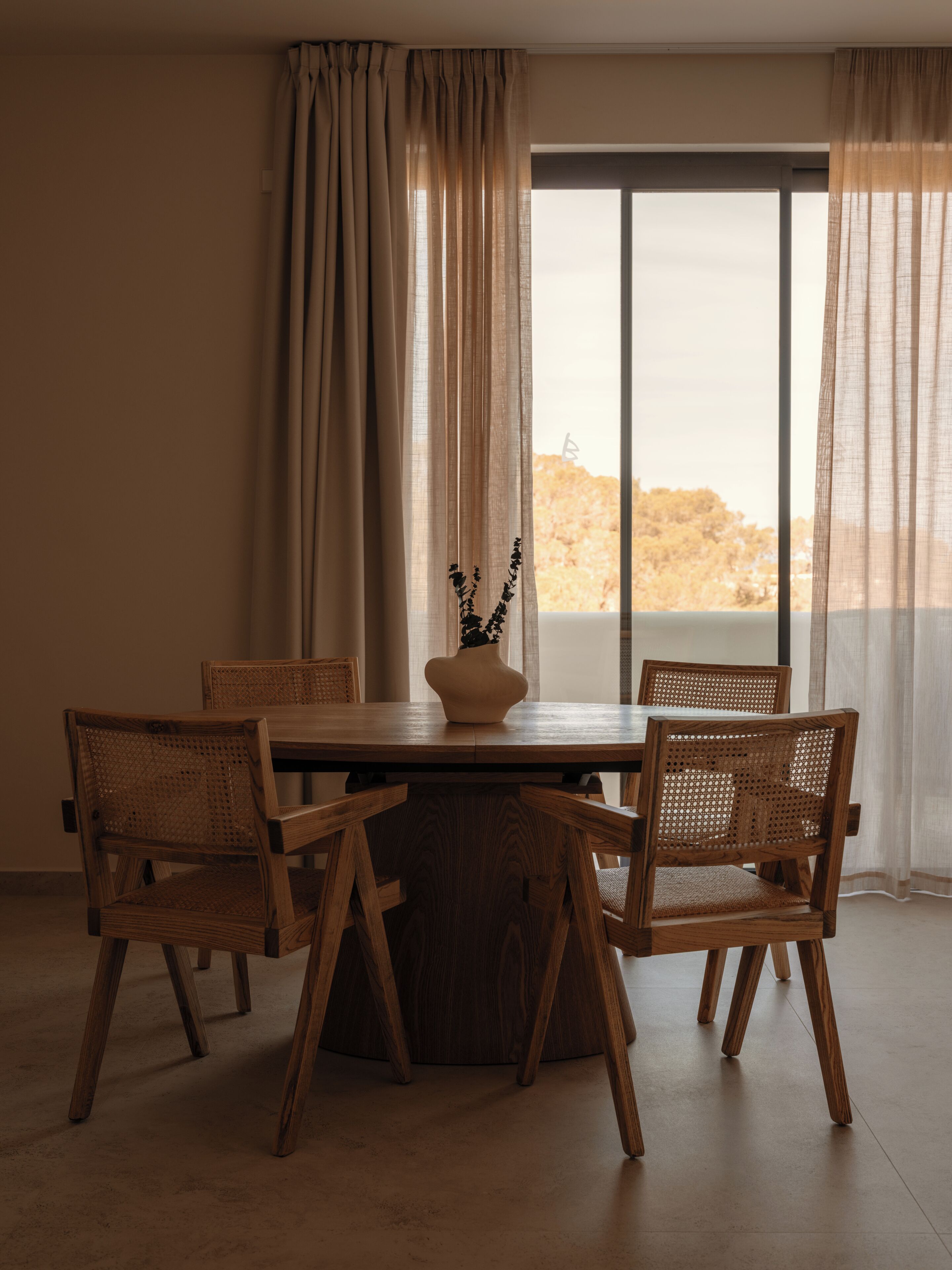 A dining area with a round wooden table, four wicker chairs, and a vase with flowers.