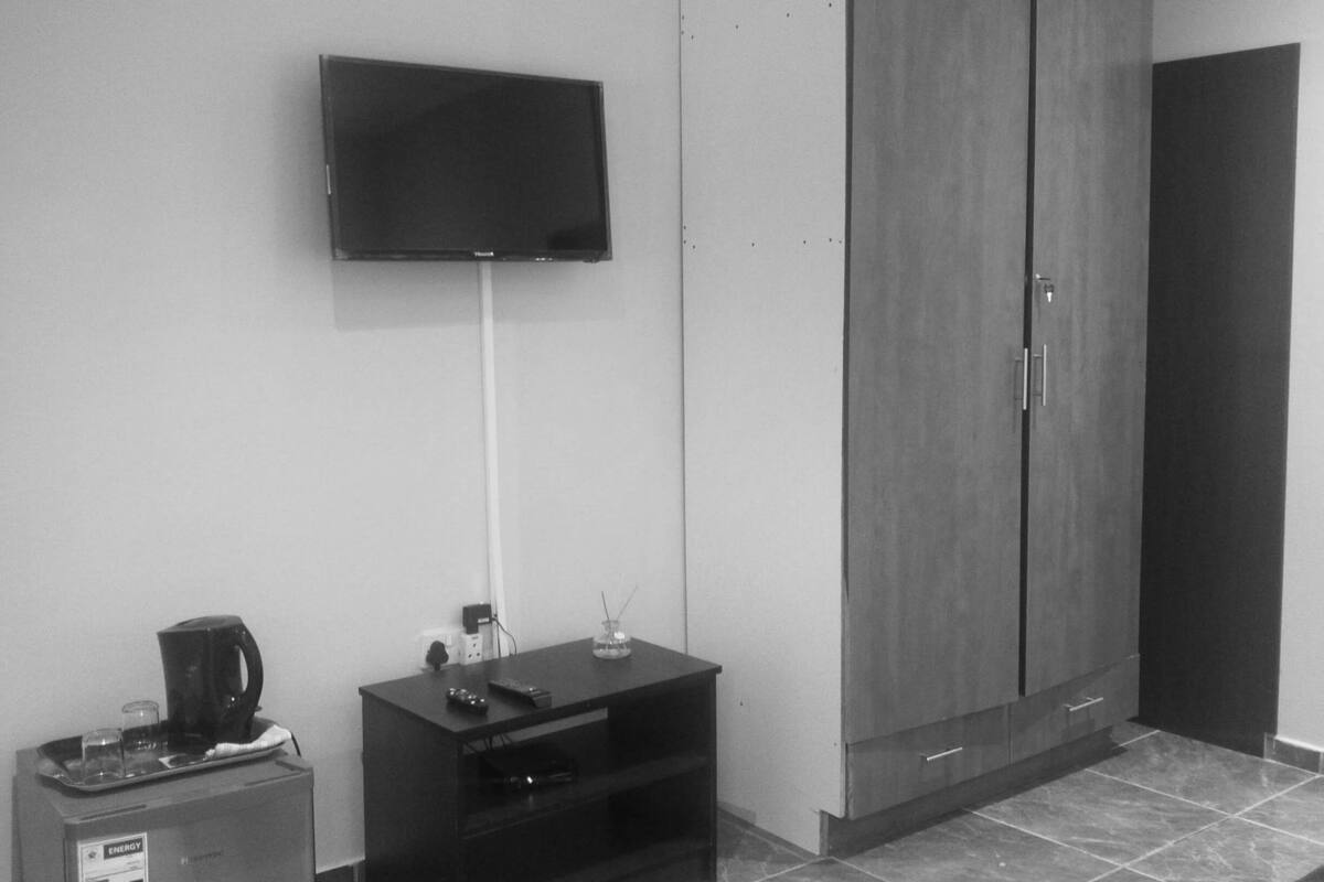 A room with a flat-screen TV mounted on the wall, a small refrigerator, a coffee maker, a cabinet, and a tiled floor.