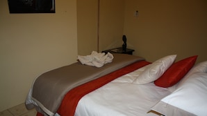 A neatly made bed with a red and white color scheme, a small bedside table with a lamp, and a wall-mounted outlet. - StayBridge Apartments Suites & Chalets (Maun)