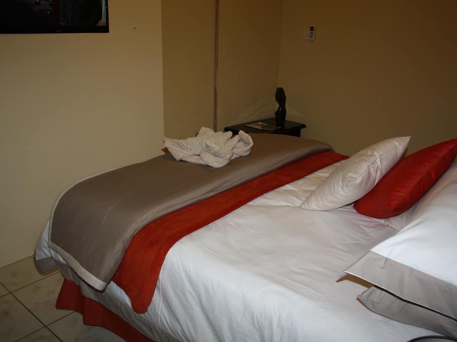 Una cama bien hecha con una decoración en rojo y blanco, una mesita de noche pequeña con una lámpara, y un enchufe en la pared.