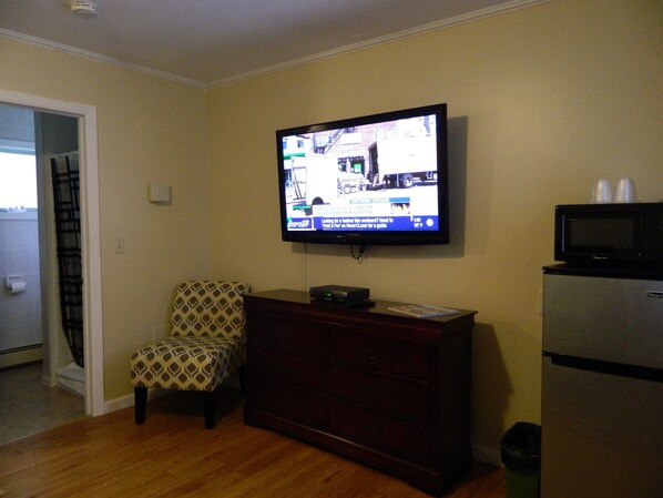 Una sala de estar con un televisor montado en la pared, un sillón estampado, un aparador de madera y un refrigerador.