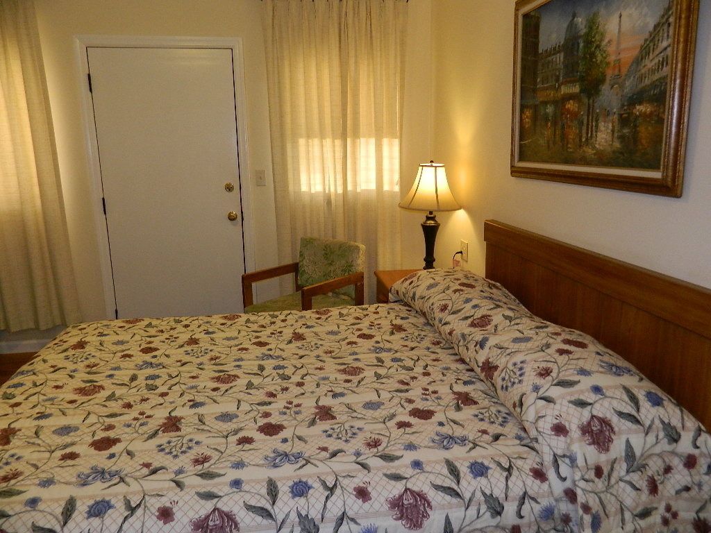 A hotel room with a floral-patterned bedspread, a wooden headboard, a chair, a lamp, and a painting on the wall.