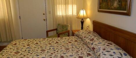 A hotel room with a floral-patterned bedspread, a wooden headboard, a chair, a lamp, and a painting on the wall.