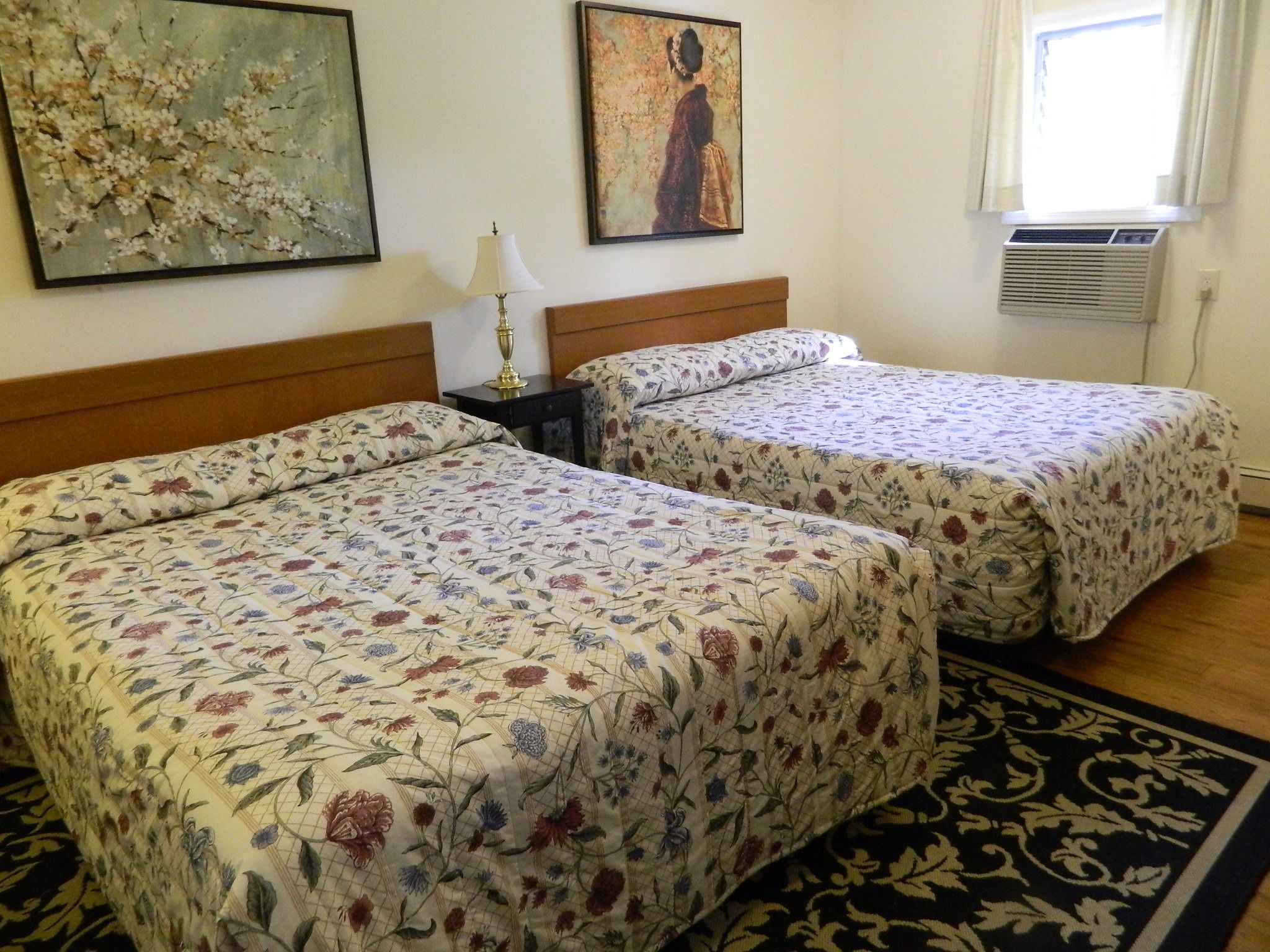 Hotelkamer met twee bedden, beddengoed met bloemenprint, een nachtkastje met een lamp, twee schilderijen aan de muur en een raam met airconditioning.