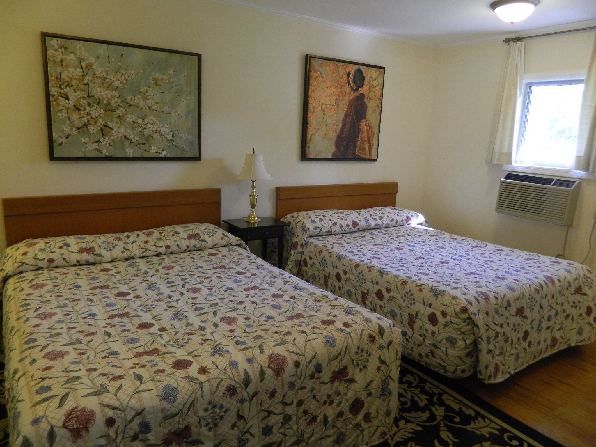 A hotel room with two beds, floral bedspreads, a nightstand with a lamp, two paintings on the wall, and an air conditioning unit.