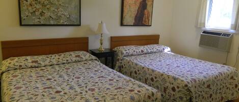 A hotel room with two beds, floral bedspreads, a nightstand with a lamp, two paintings on the wall, and an air conditioning unit.