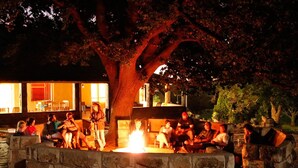 A group of people gathered around a fire pit in an outdoor setting with stone seating and a large tree. - Imla Guest Farm (Setsoto)