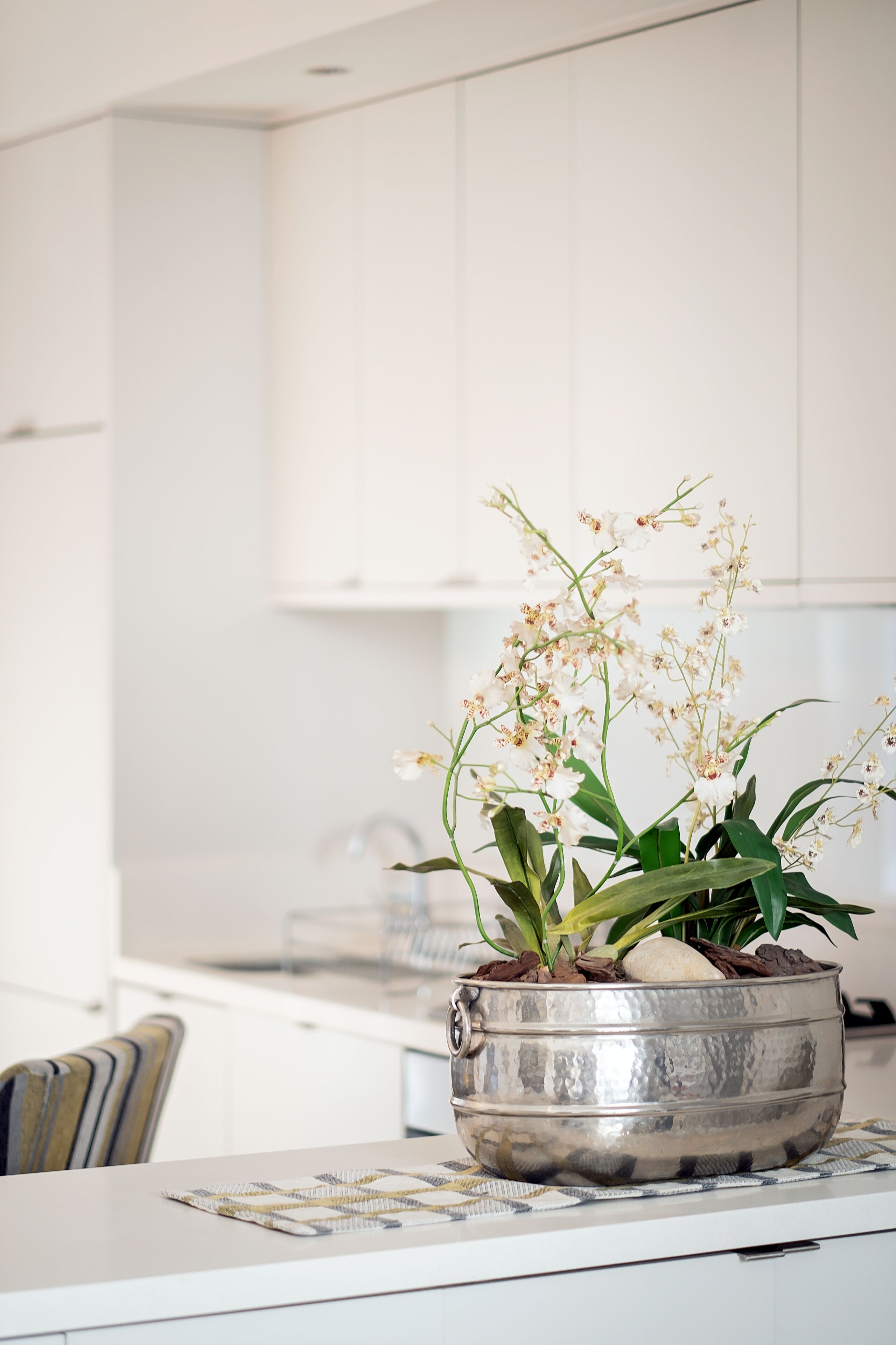 Une cuisine avec des meubles blancs, une casserole en acier inoxydable ornée d’orchidées et un set de table à motifs posé sur le comptoir.