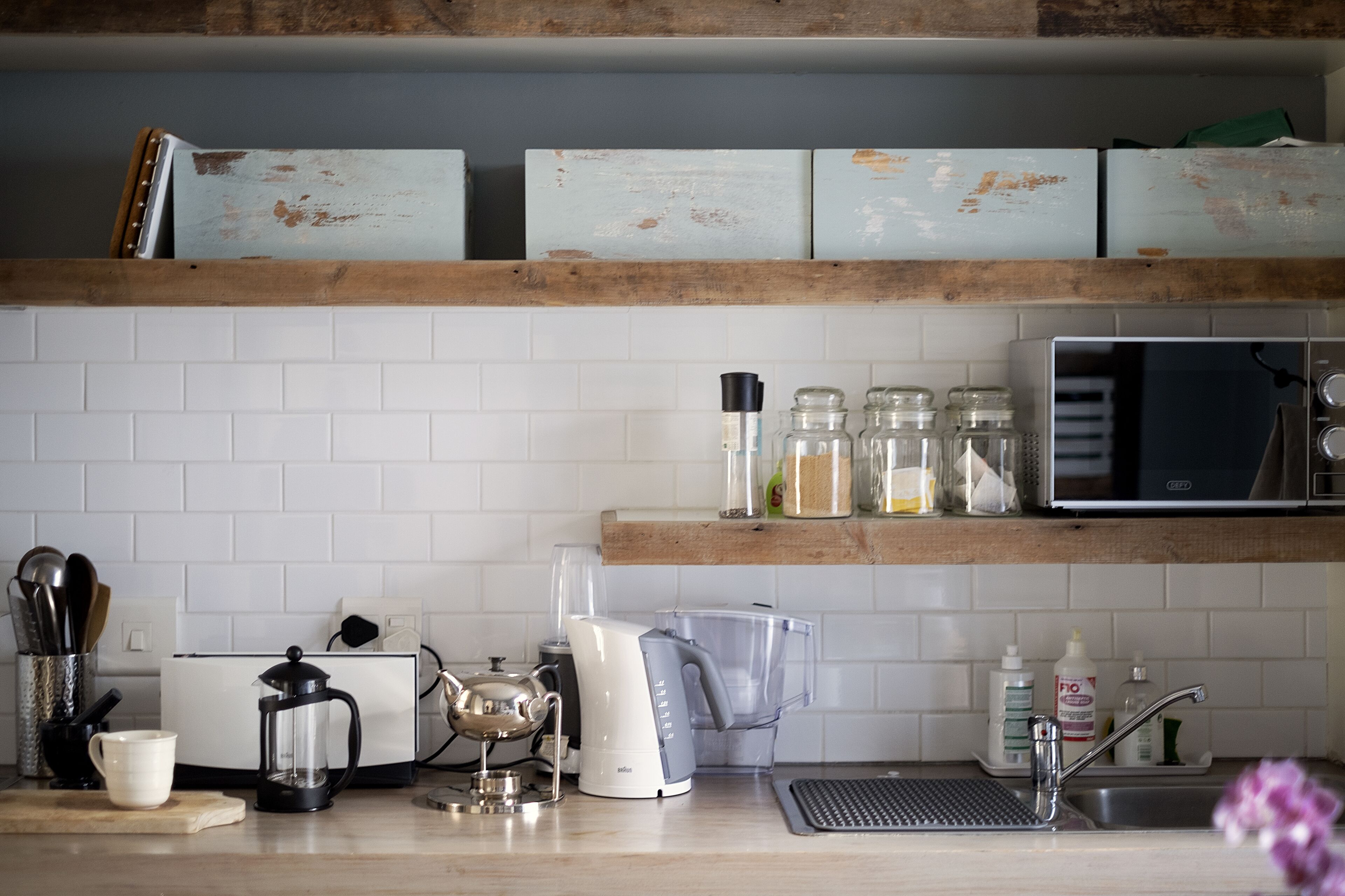 Une cuisine avec des carreaux blancs, une machine à café, une bouilloire, un mixeur et un four à micro-ondes placés sur une étagère en bois.