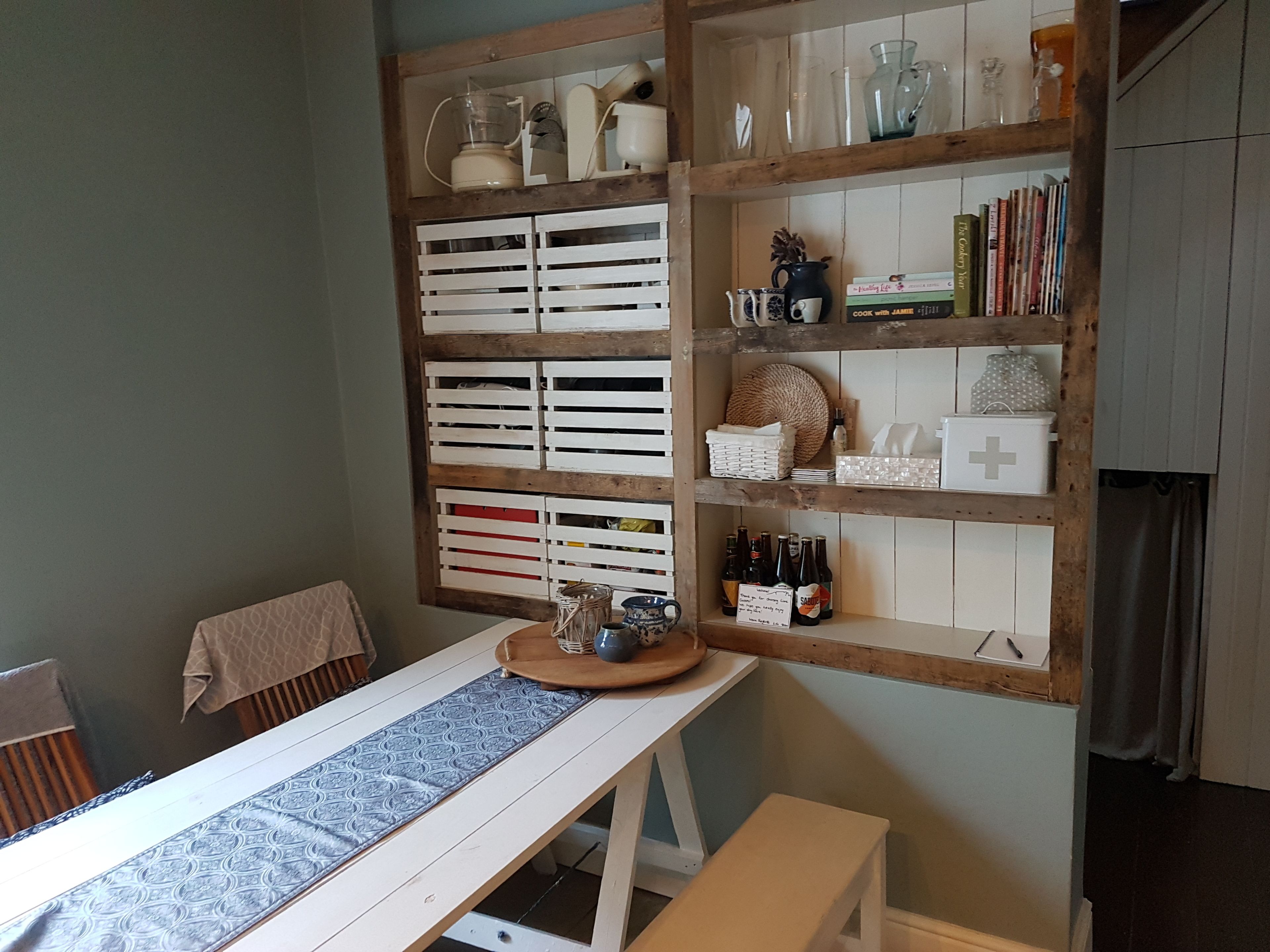 A kitchen with a wooden shelf unit containing kitchenware, a table with a patterned runner, and a bench.