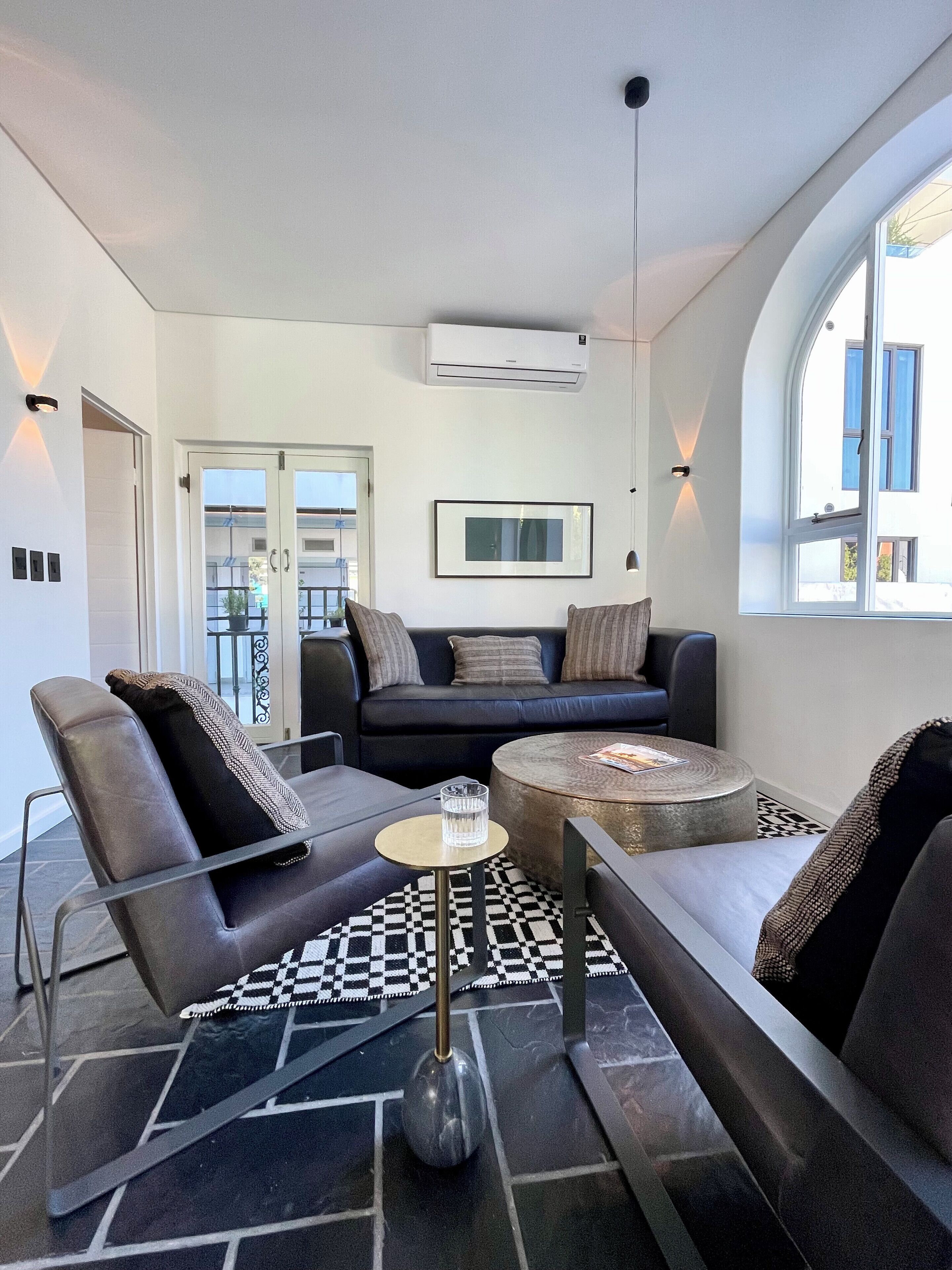 A modern living room with a black and white checkered floor, a round coffee table, a black leather sofa, and a black leather armchair.