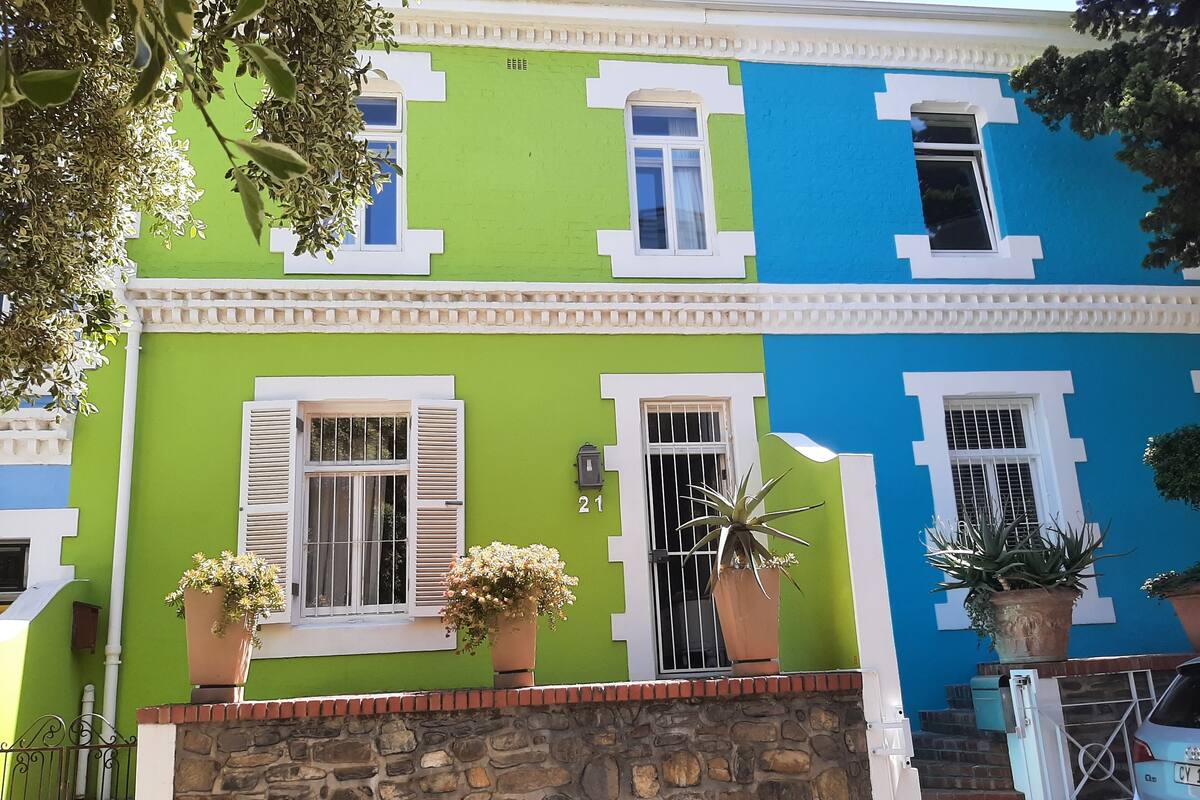 A colorful building with three distinct sections, each painted in a different color. There are potted plants on the front porch, a stone wall, and a metal gate leading to the entrance.