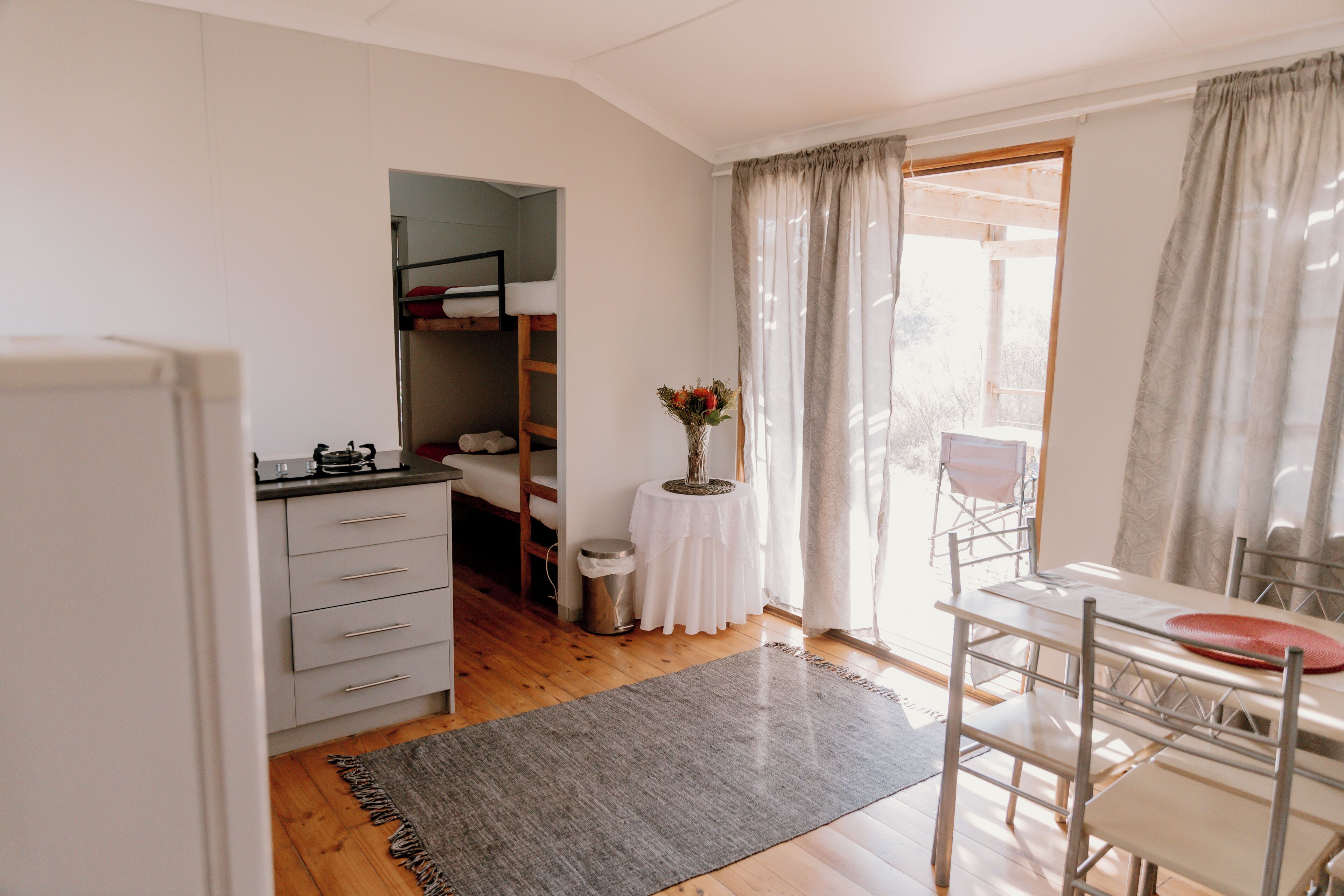 A compact kitchen with wooden cabinets, a dining area with a table and chairs, a small round table with a vase, and a view through a window with curtains.