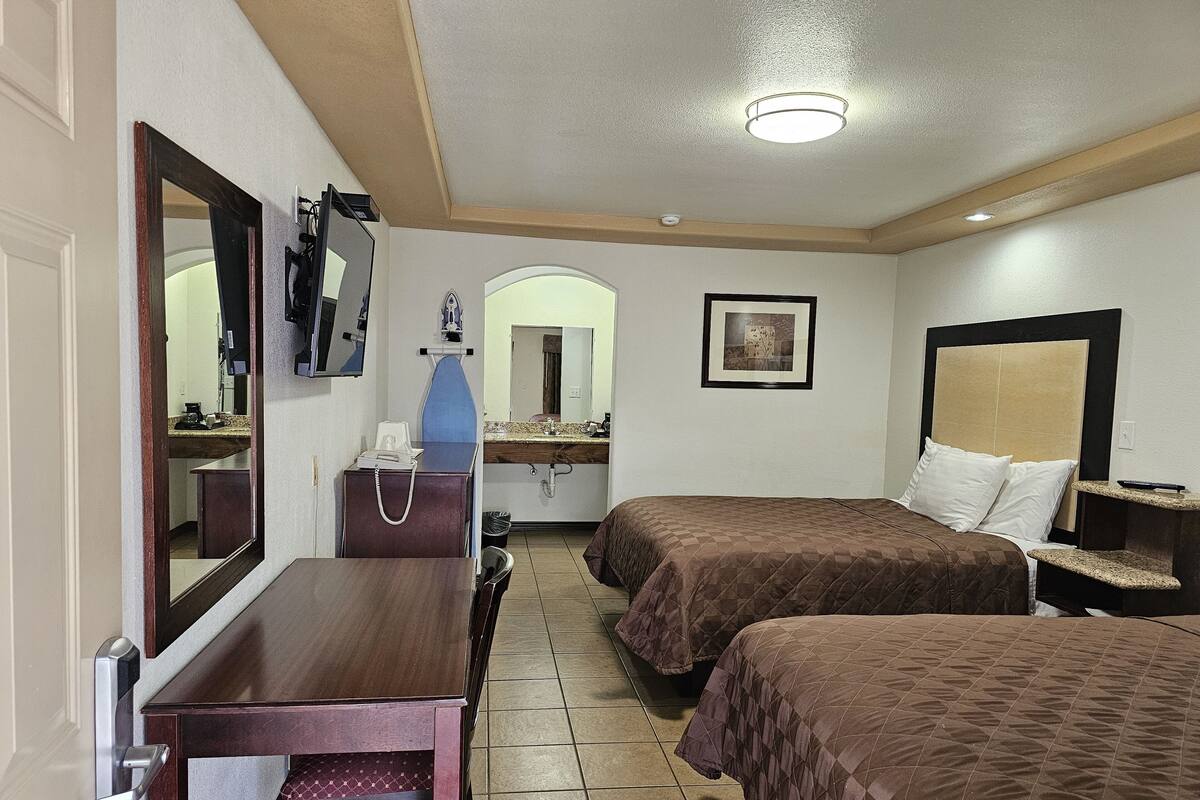 A hotel room with two beds, a desk, a dresser, a mirror, a sink, and a framed picture on the wall.