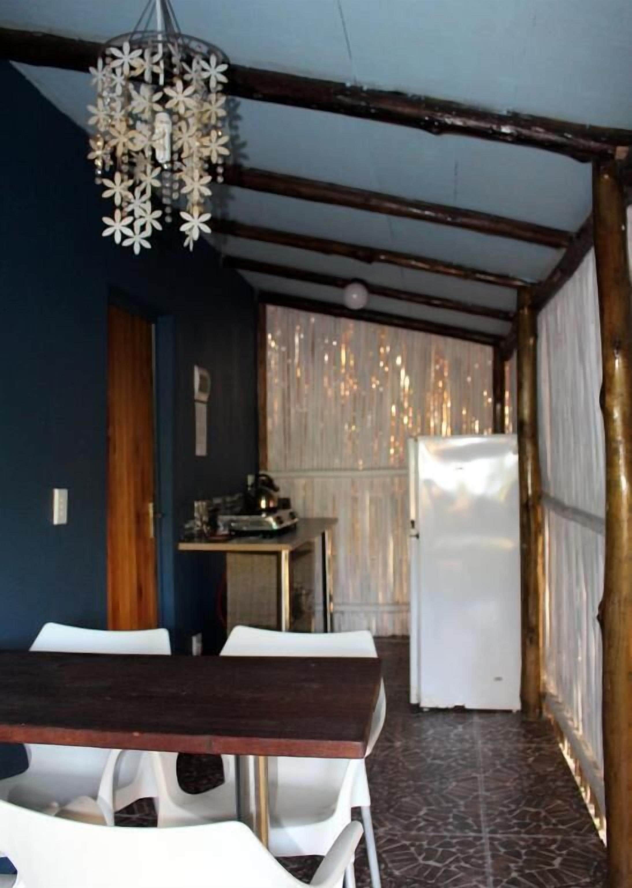 Un comedor con mesa y sillas de madera, una lámpara colgante y una zona de cocina con refrigerador.