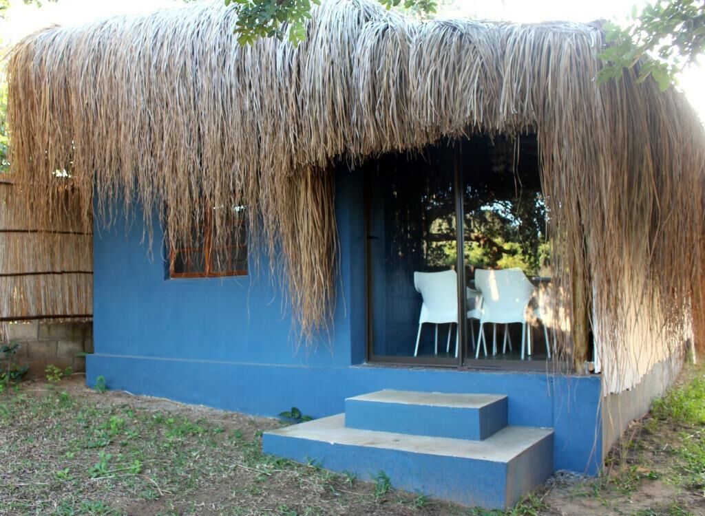 A thatched-roof structure with a blue exterior, a small set of steps leading to a door, and a table and chairs outside.