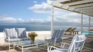 A terrace with a glass table and striped chairs, overlooking a pool and the sea.