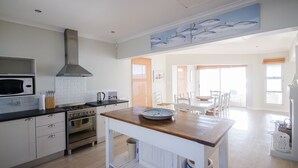 A modern kitchen with white cabinetry, a wooden countertop, and a stainless steel range hood. There is a dining area with white chairs and a table, and a large window providing natural light.