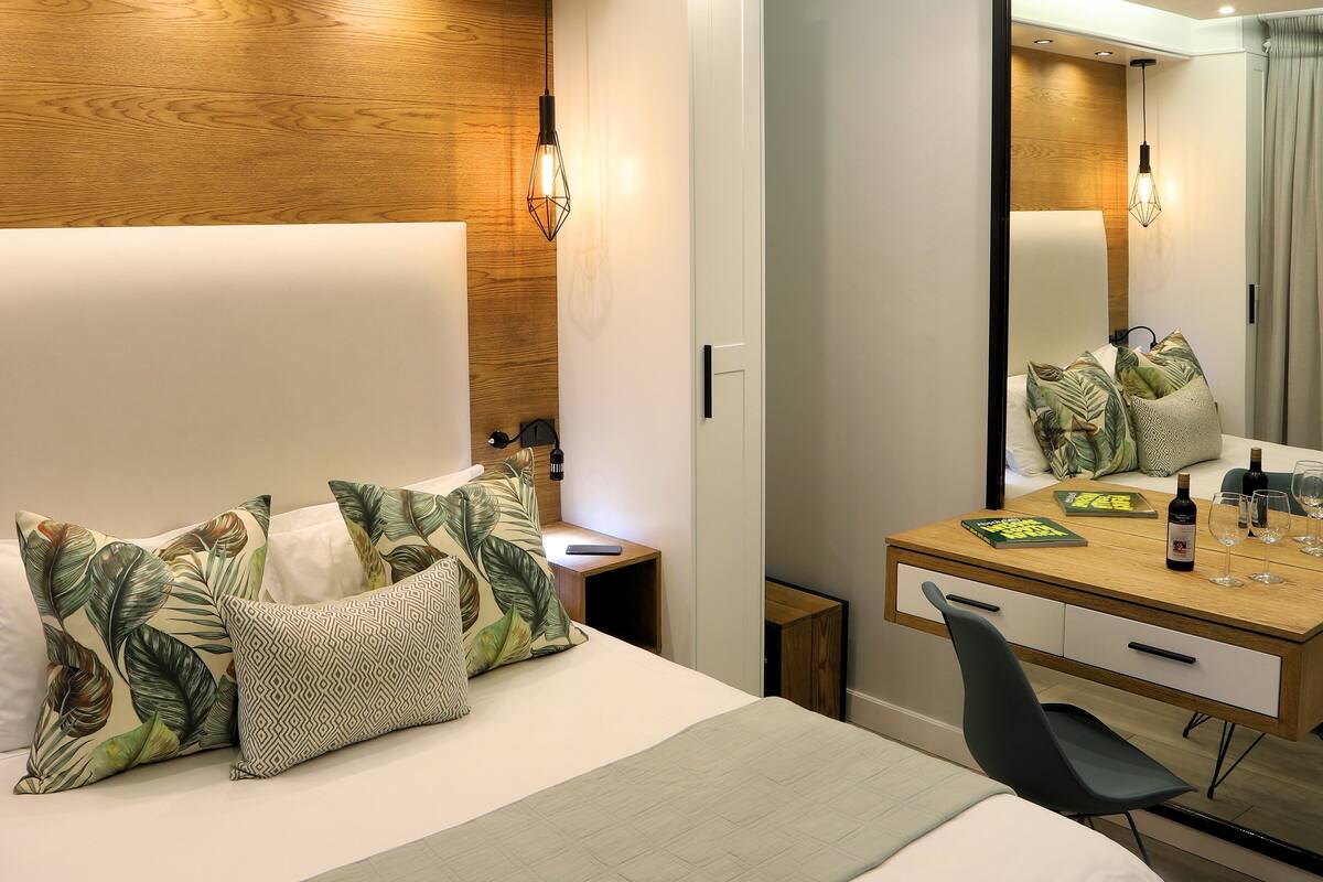 A modern hotel room with a large bed, a wooden accent wall, a desk with a mirror, and a chair.
