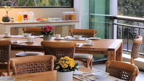 Un espace repas avec des tables en bois et des chaises en osier, une horloge au mur et une vue sur la verdure à l’extérieur.