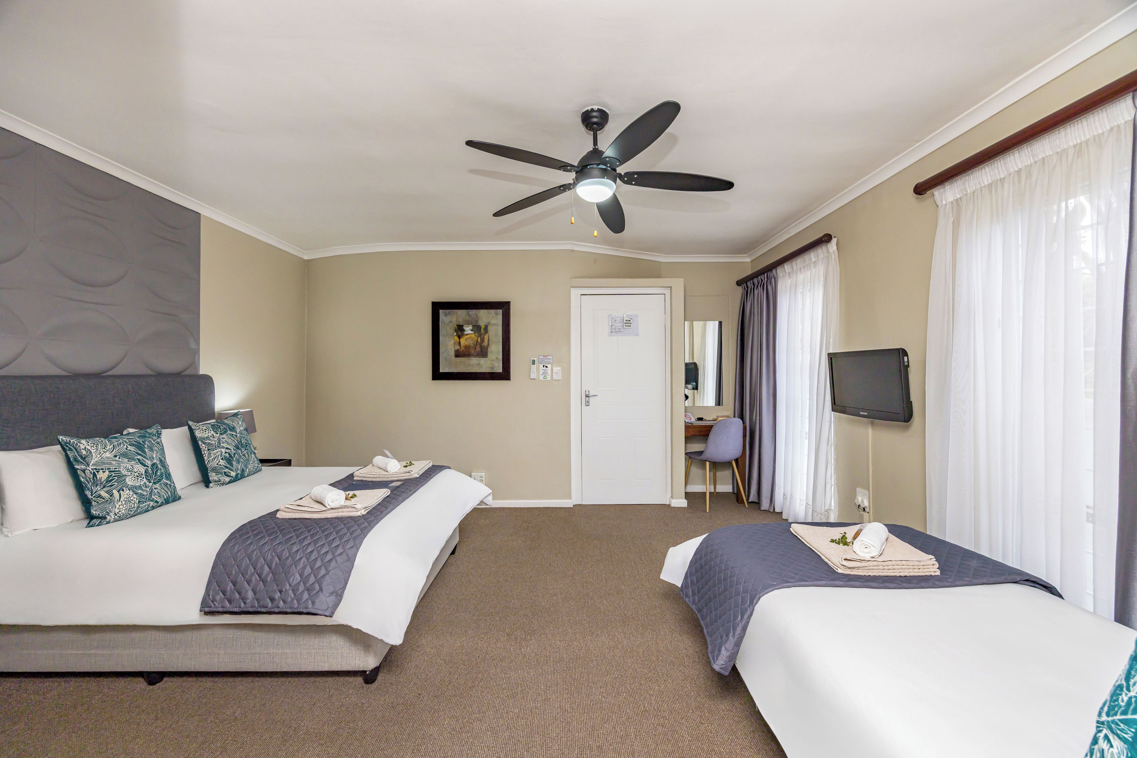 A hotel room with two beds, a ceiling fan, a desk with a chair, a TV, and a door.