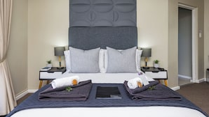 Un lit bien fait, recouvert d’une couverture sombre, avec deux oreillers et une serviette pliée de chaque côté. On y trouve deux tables de chevet avec des lampes et de petites plantes.