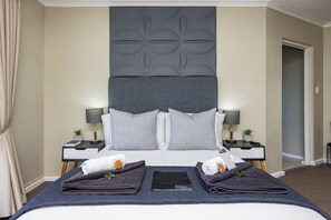 Un lit bien fait, recouvert d’une couverture sombre, avec deux oreillers et une serviette pliée de chaque côté. On y trouve deux tables de chevet avec des lampes et de petites plantes.