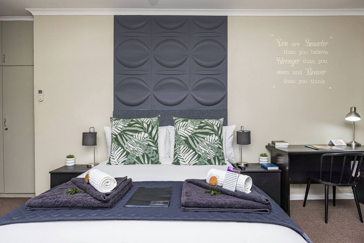 Een moderne hotelkamer met een bed, een bureau, een stoel en een inspirerende quote op de muur.