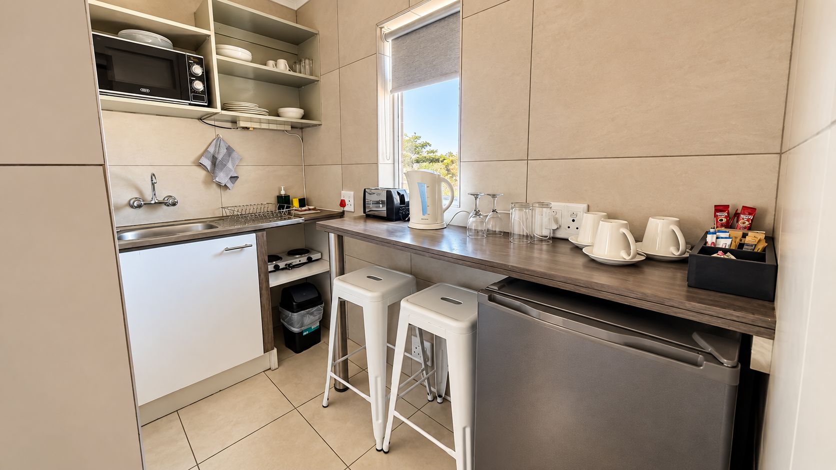 A compact kitchen with a microwave, sink, and stainless steel refrigerator. There are two white bar stools and a trash bin.