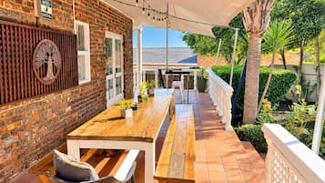 An outdoor patio with a wooden table and benches, surrounded by a brick wall and a white fence.
