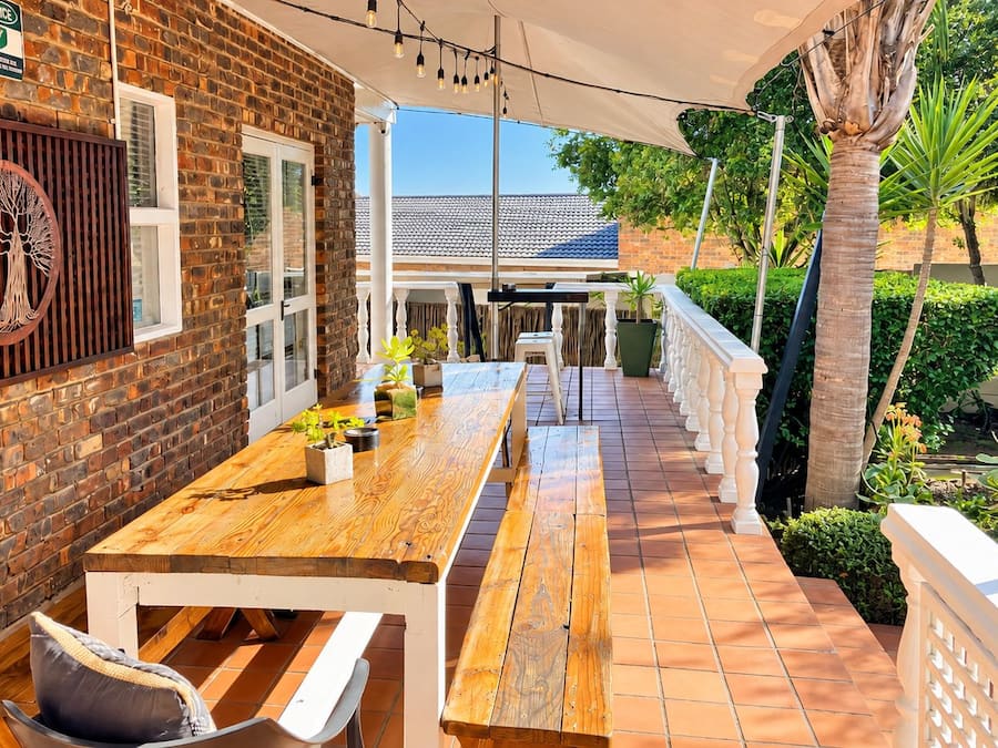 Un patio al aire libre con una mesa y bancos de madera, rodeado por un muro de ladrillo y una valla blanca.