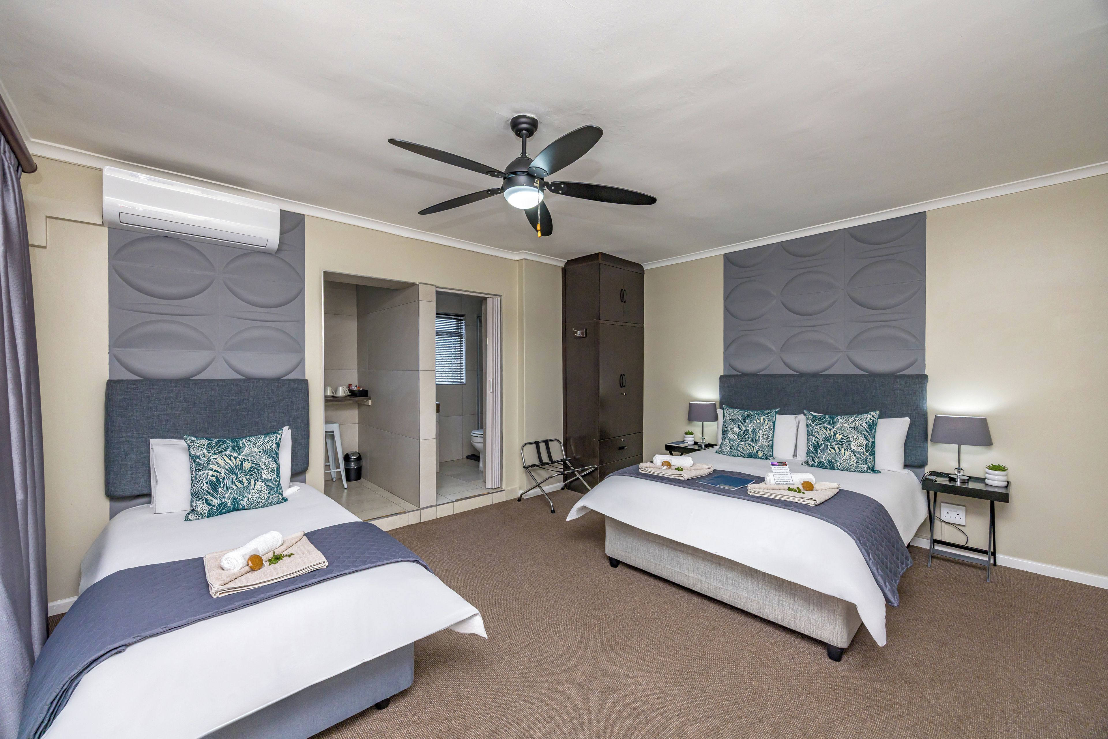 Une chambre d’hôtel avec deux lits, un ventilateur de plafond, un minibar et une salle de bain visible à travers une porte ouverte.