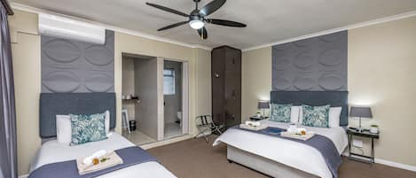 Habitación de hotel con dos camas, ventilador de techo, minibar y baño visible a través de una puerta abierta.