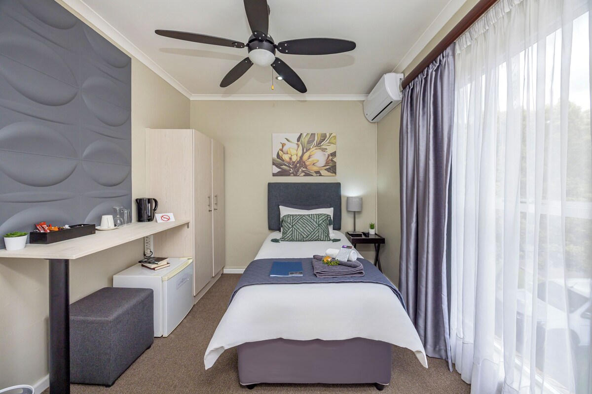 Une chambre d’hôtel équipée d’un lit, d’un bureau, d’une chaise, d’un ventilateur de plafond, d’une télévision et d’une fenêtre avec des rideaux.
