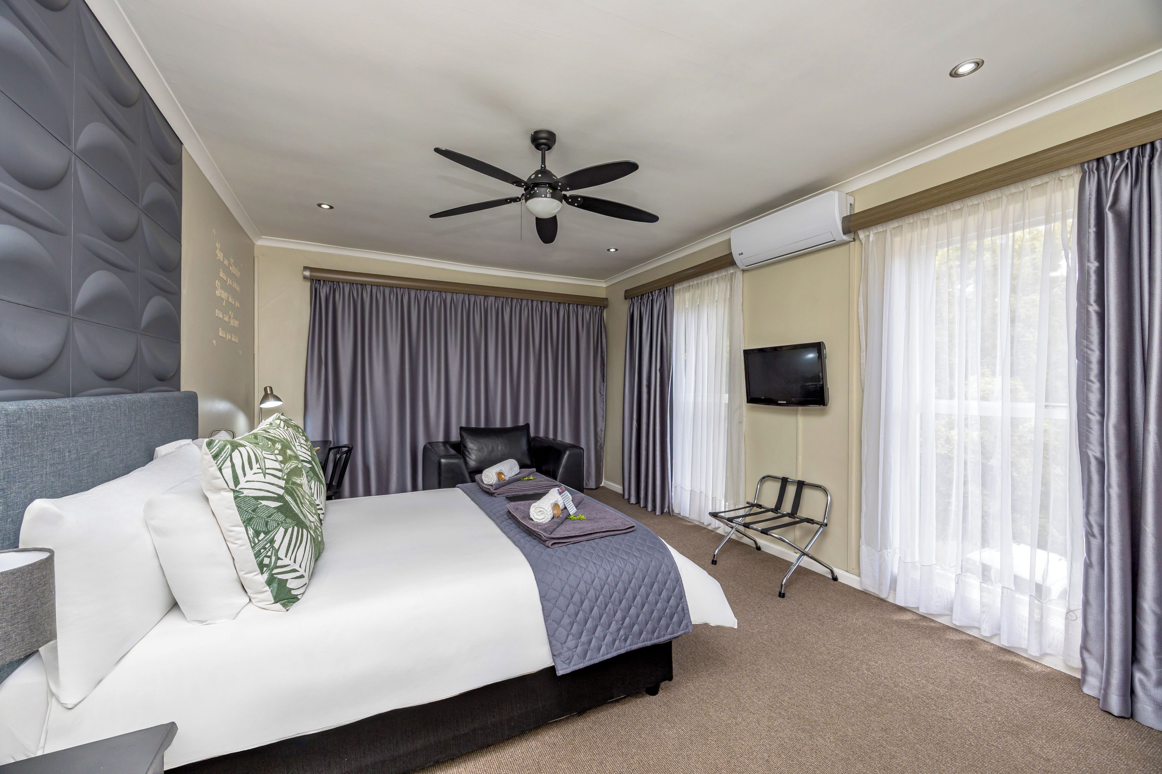 A hotel room with a bed, a ceiling fan, a TV, and a window with curtains.