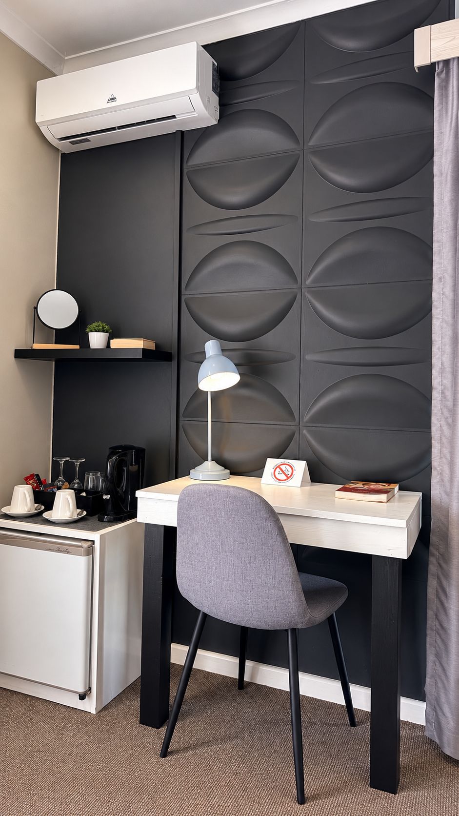 Habitación de hotel compacta con escritorio, silla, equipo de aire acondicionado y un panel decorativo en la pared.