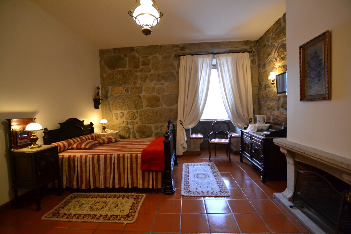A bedroom with a stone wall, a bed with striped bedding, a fireplace, and a nightstand with a lamp.