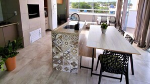 A modern kitchen with a built-in oven, a dining area with a long table and chairs, and a view of the city through large windows. - Apartamento do Sol (Bombinhas)