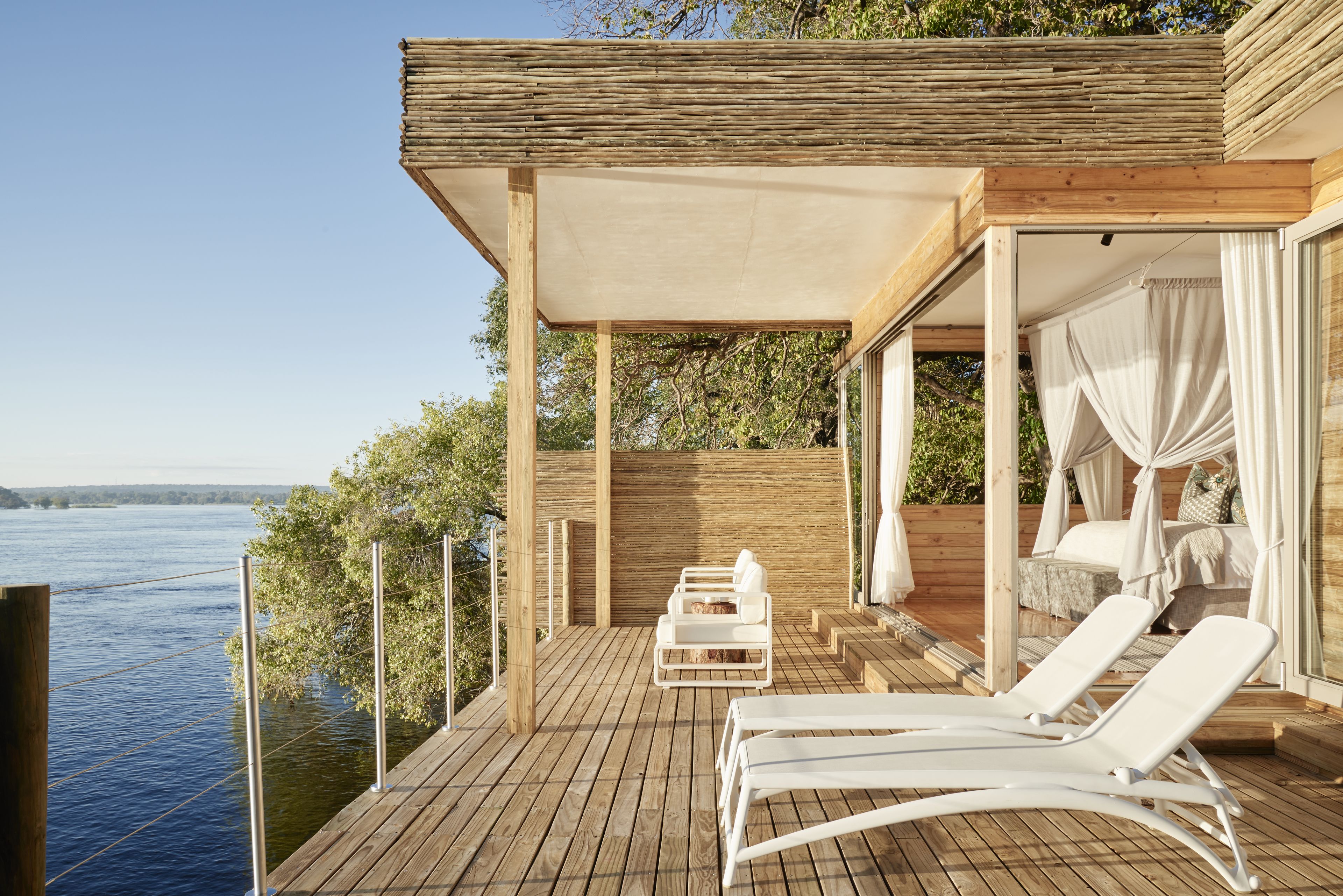 A wooden deck with white lounge chairs and a canopy bed overlooking a lake.