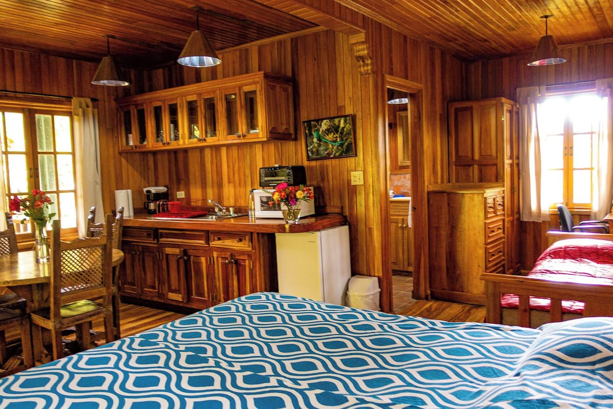 A wooden cabin interior with a bed, a dining table, a kitchen area, and a window.