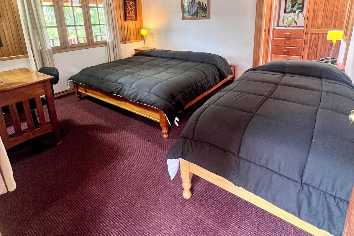 A room with two beds, wooden ceiling beams, a window with curtains, a small table, and a wall-mounted lamp.
